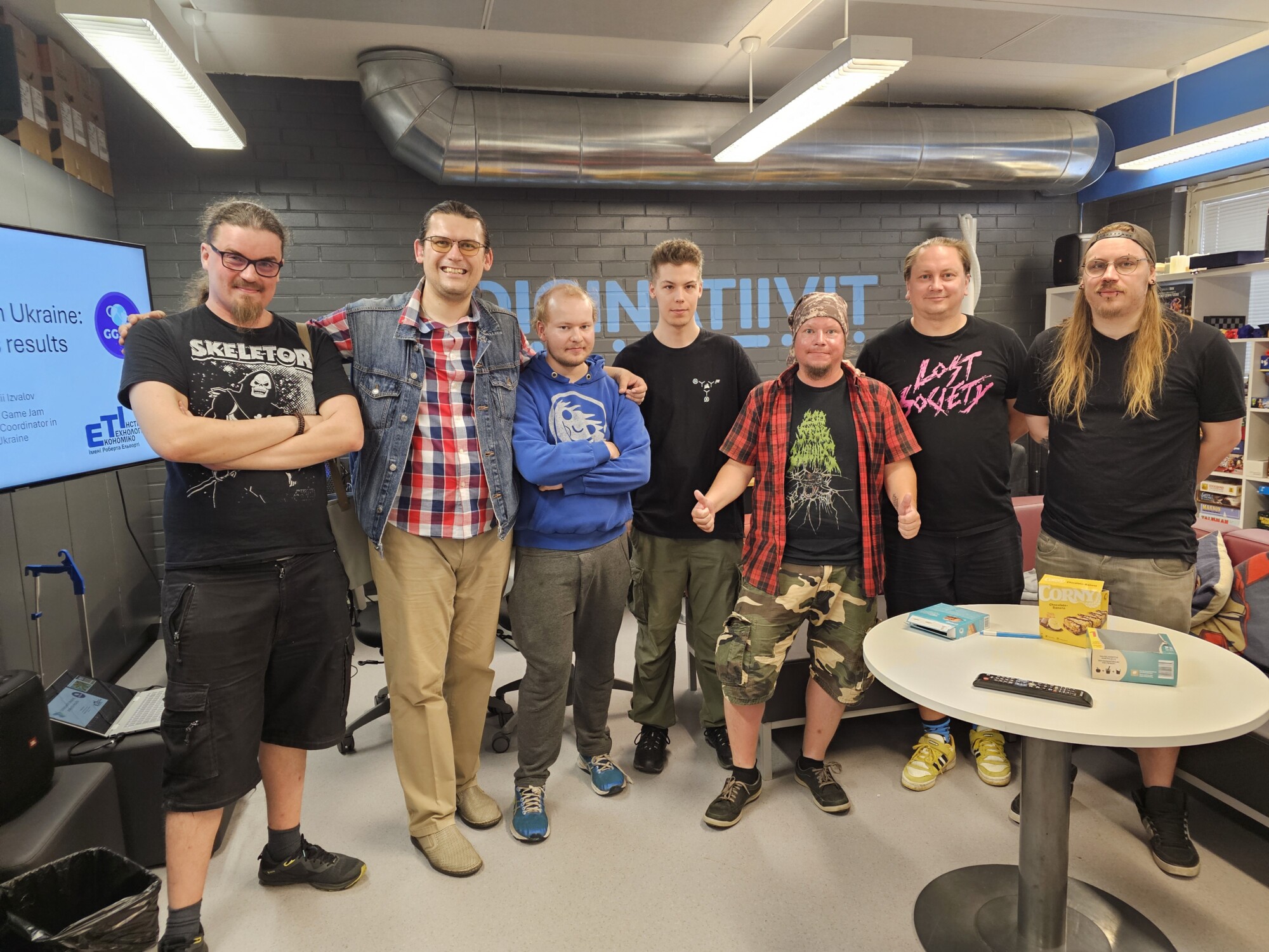 Seitsemän ihmistä seisoo vierekkäin rennossa sisäympäristössä ja hymyilee kameralle. He ovat pukeutuneet rentoihin vaatteisiin, joissa on graafisia t-paitoja, ruudullisia paitoja ja huppareita. Taustalla on teknisiä laitteita ja suuri näyttö.