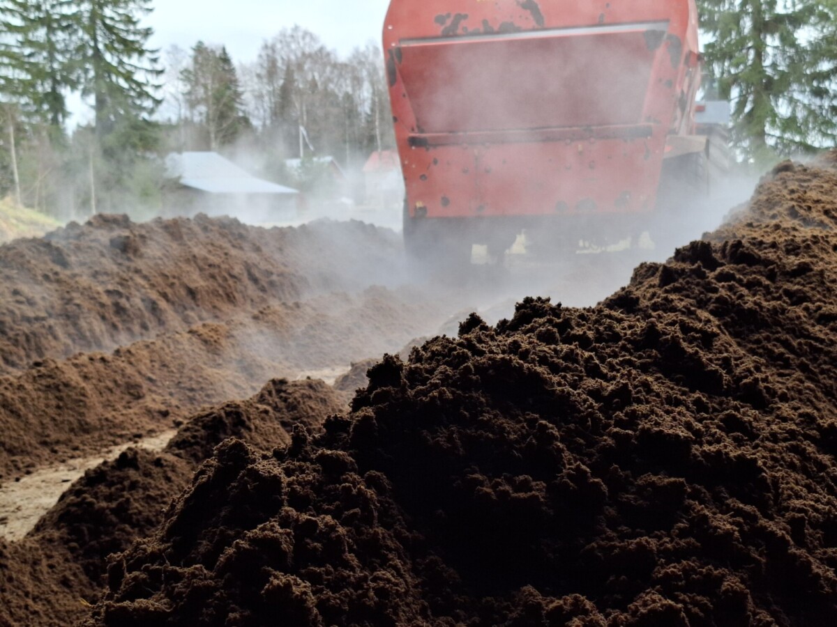 Punainen kone levittää höyryävää kompostia tai multaa suuriin kasoihin ulkona, ja taustalla näkyy puita ja taloja pilvisenä päivänä.