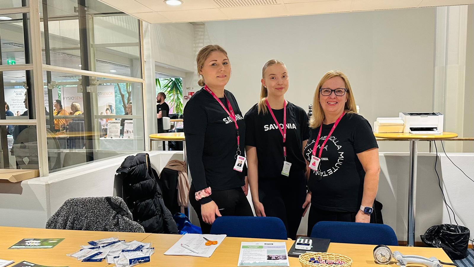 Kolme naista seisoo ja hymyilee infopöydän takana, jossa on esitteitä, esitteitä ja pieniä esineitä. Kaikilla on mustat Savonia-paidat ja nimikyltit. Paikka vaikuttaa olevan sisätiloissa järjestettävä yleisötapahtuma tai näyttely.