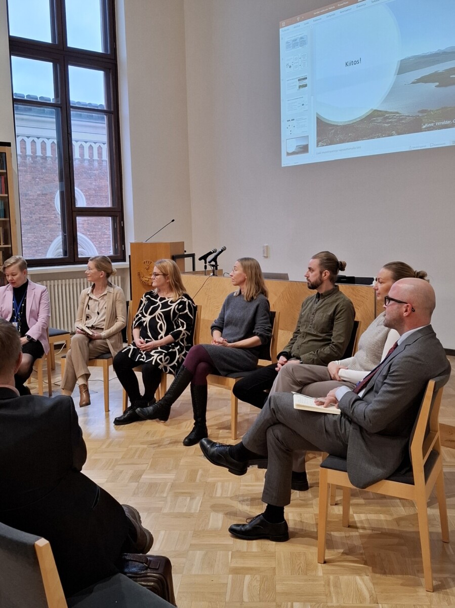 Seitsemän ihmistä istuu tuoleilla paneelikeskustelussa valoisassa huoneessa, jossa on puulattia. Taustalla näkyy suuri ikkuna ja näyttö, jossa näkyy esitys.