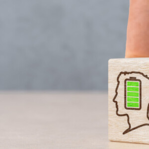 Käsi pyörittää puupalikkaa, jossa on kaksi ihmisprofiilia: toisessa on täysi vihreä akku ja toisessa lähes tyhjä punainen akku, jotka symboloivat henkistä tai energiatasoa.