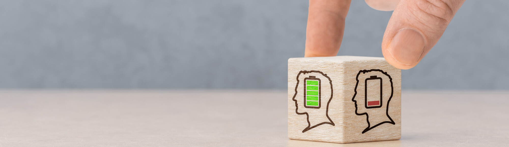 Käsi pyörittää puupalikkaa, jossa on kaksi ihmisprofiilia: toisessa on täysi vihreä akku ja toisessa lähes tyhjä punainen akku, jotka symboloivat henkistä tai energiatasoa.