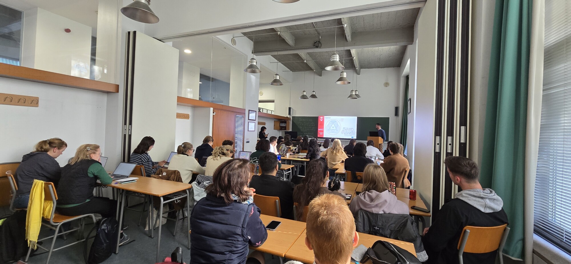 Opiskelijaryhmä istuu luokkahuoneessa luennoitsijaa vastapäätä, joka esittää dioja huoneen etuosassa olevalla näytöllä. Huone on hyvin valaistu, ja siinä on suuret ikkunat ja modernit kalusteet.