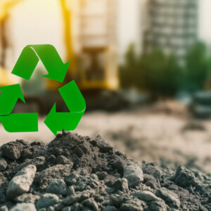 Vihreä kierrätyssymboli seisoo rakennustyömaan maakasan päällä, ja taustalla on epäselviä kaivinkoneita ja rakennuksia, jotka edustavat kierrätystä tai kestävyyttä rakentamisessa.