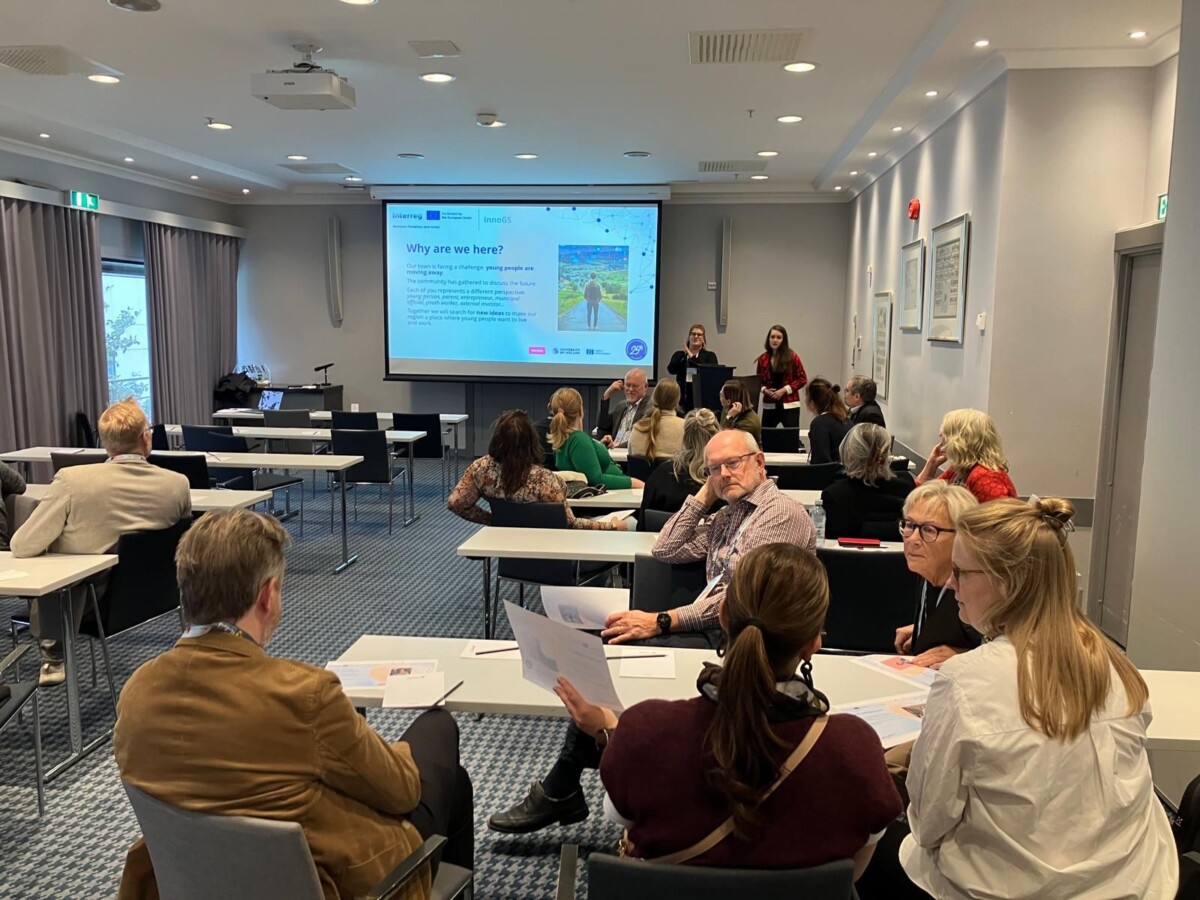 Ihmiset istuvat neuvotteluhuoneen pöydissä ja keskustelevat, kun edessä olevalla suurella näytöllä näkyy esitys. Useat esittelijät seisovat näytön lähellä ja puhuvat ryhmälle.