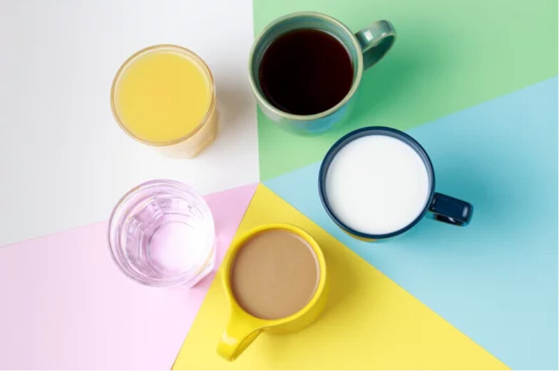 Viisi kuppia, jotka sisältävät erilaisia juomia - appelsiinimehua, mustaa kahvia, maitoa, vettä ja maitokahvia - on sijoitettu pastellinväriselle, geometriselle taustalle ylhäältä katsottuna.