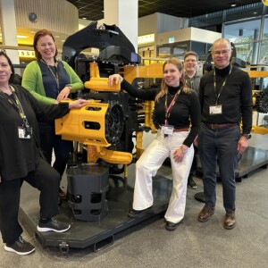 Viisi ihmistä seisoo suuren keltaisen teollisuuskoneen ympärillä sisätiloissa ja hymyilee kameralle. Ympäristö näyttää olevan moderni esittely- tai näyttelytila, jonka taustalla on lasiseinät ja kylttejä.