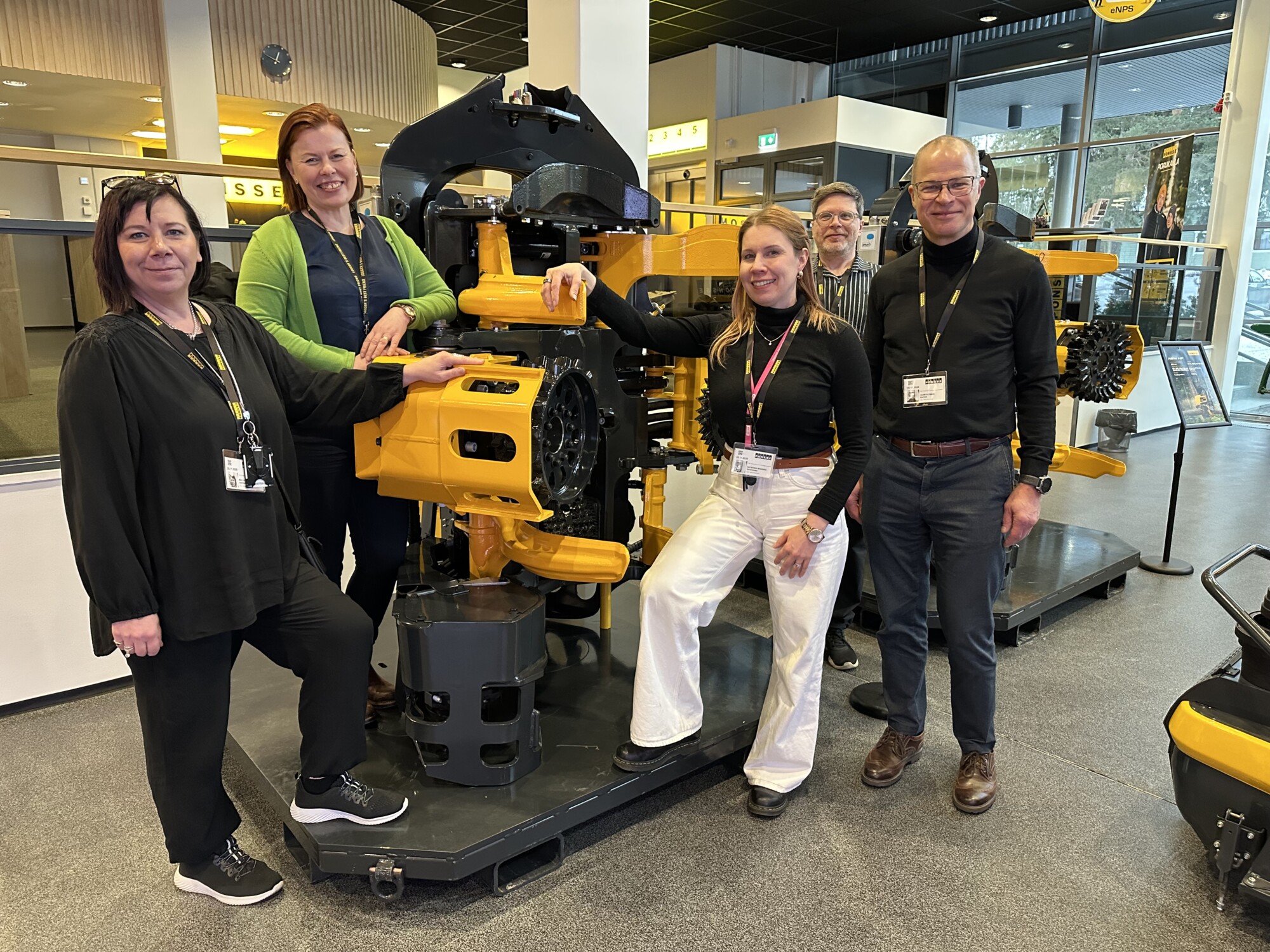 Viisi ihmistä seisoo suuren keltaisen teollisuuskoneen ympärillä sisätiloissa ja hymyilee kameralle. Ympäristö näyttää olevan moderni esittely- tai näyttelytila, jonka taustalla on lasiseinät ja kylttejä.