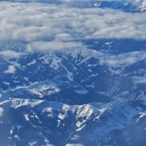 Ilmakuva lumen peittämistä vuorenhuipuista, jotka ovat osittain pilvien peitossa ja luovat dramaattisen ja luonnonkauniin maiseman.