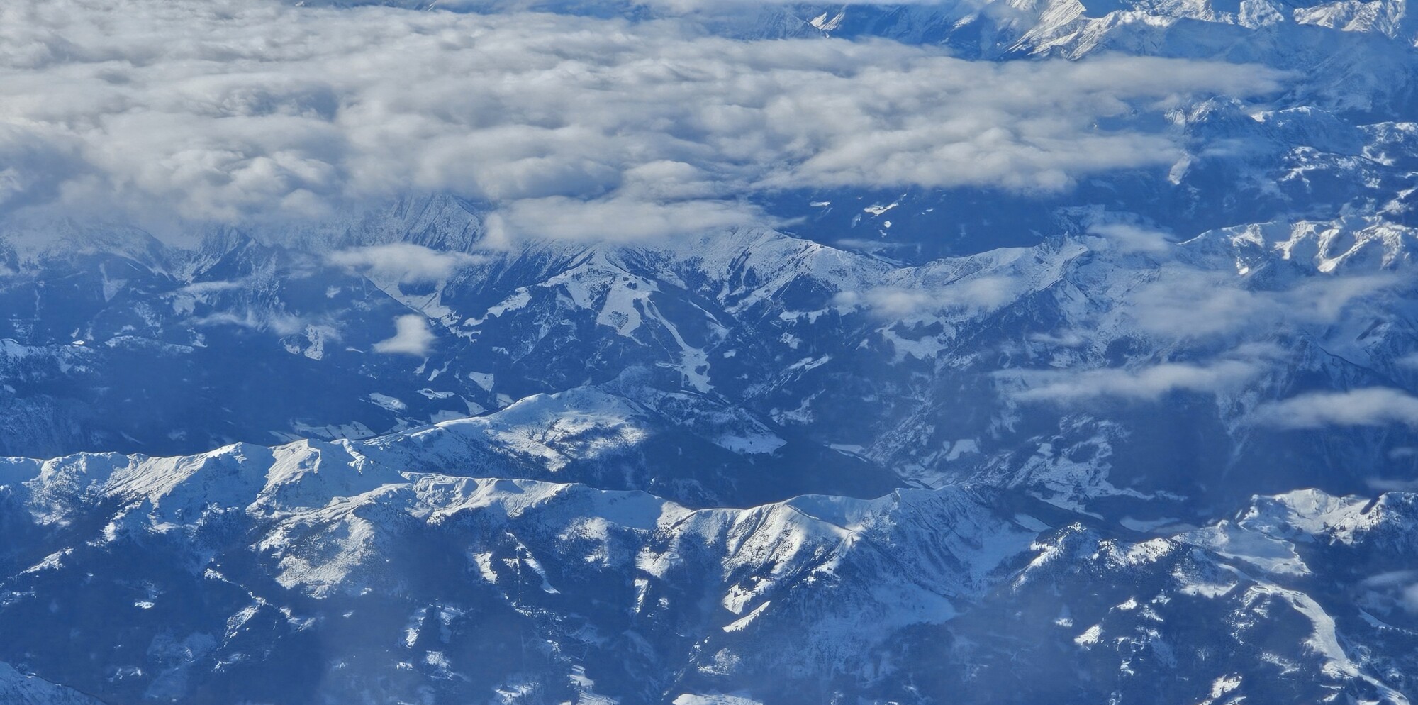 Ilmakuva lumen peittämistä vuorenhuipuista, jotka ovat osittain pilvien peitossa ja luovat dramaattisen ja luonnonkauniin maiseman.