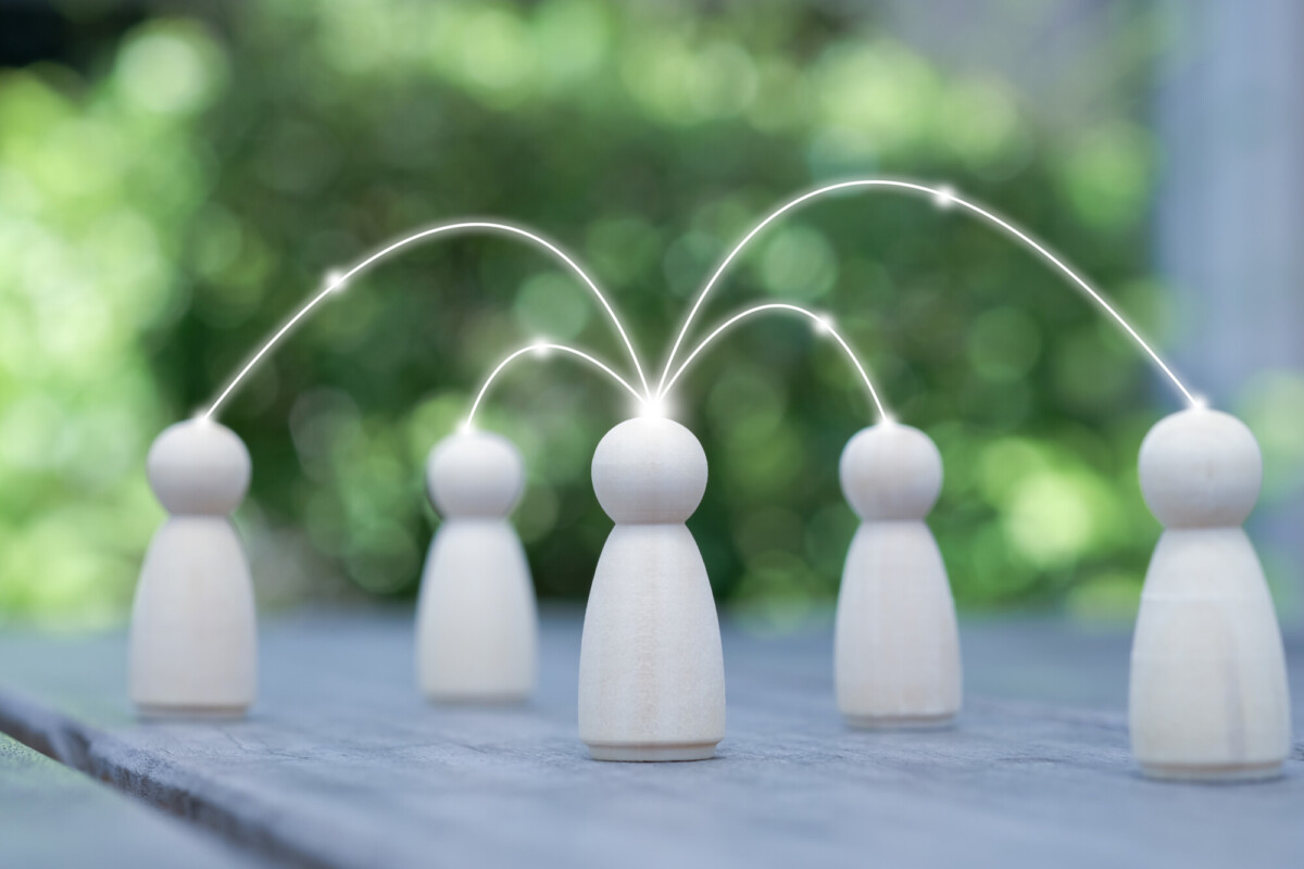 Neljä puista hahmoa on sijoitettu pöydälle ulkona, ja hehkuvat viivat yhdistävät yhden keskushahmon muihin hahmoihin, mikä symboloi viestintää tai verkostoitumista. Tausta on sumea ja vihreä.