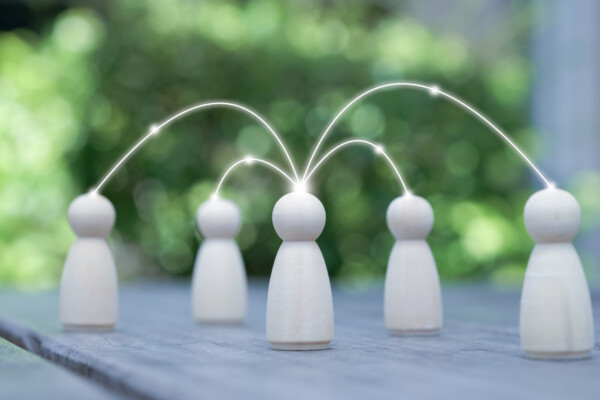 Neljä puista hahmoa on sijoitettu pöydälle ulkona, ja hehkuvat viivat yhdistävät yhden keskushahmon muihin hahmoihin, mikä symboloi viestintää tai verkostoitumista. Tausta on sumea ja vihreä.