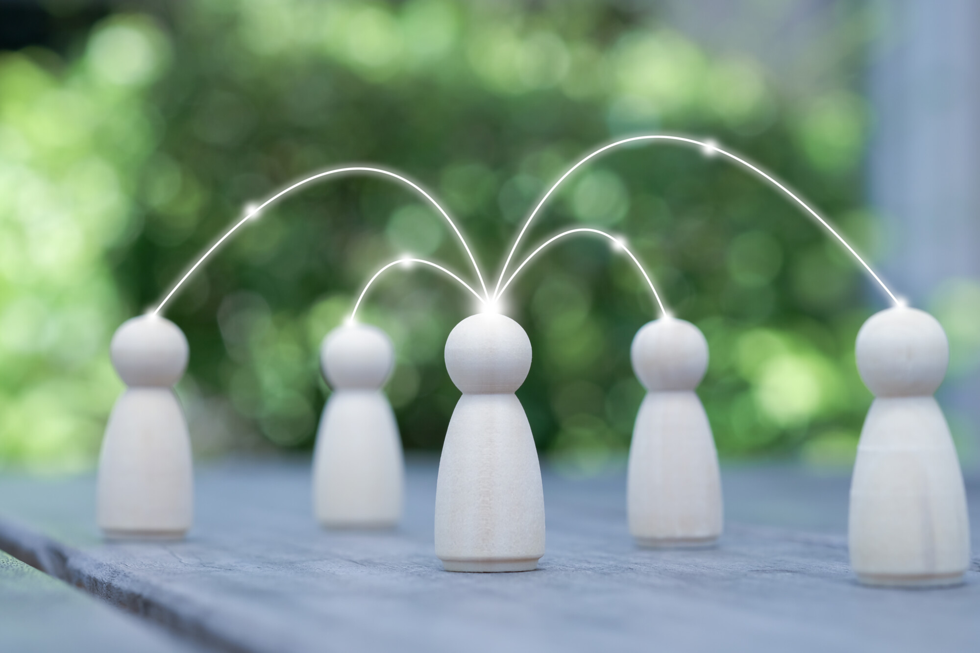 Neljä puista hahmoa on sijoitettu pöydälle ulkona, ja hehkuvat viivat yhdistävät yhden keskushahmon muihin hahmoihin, mikä symboloi viestintää tai verkostoitumista. Tausta on sumea ja vihreä.