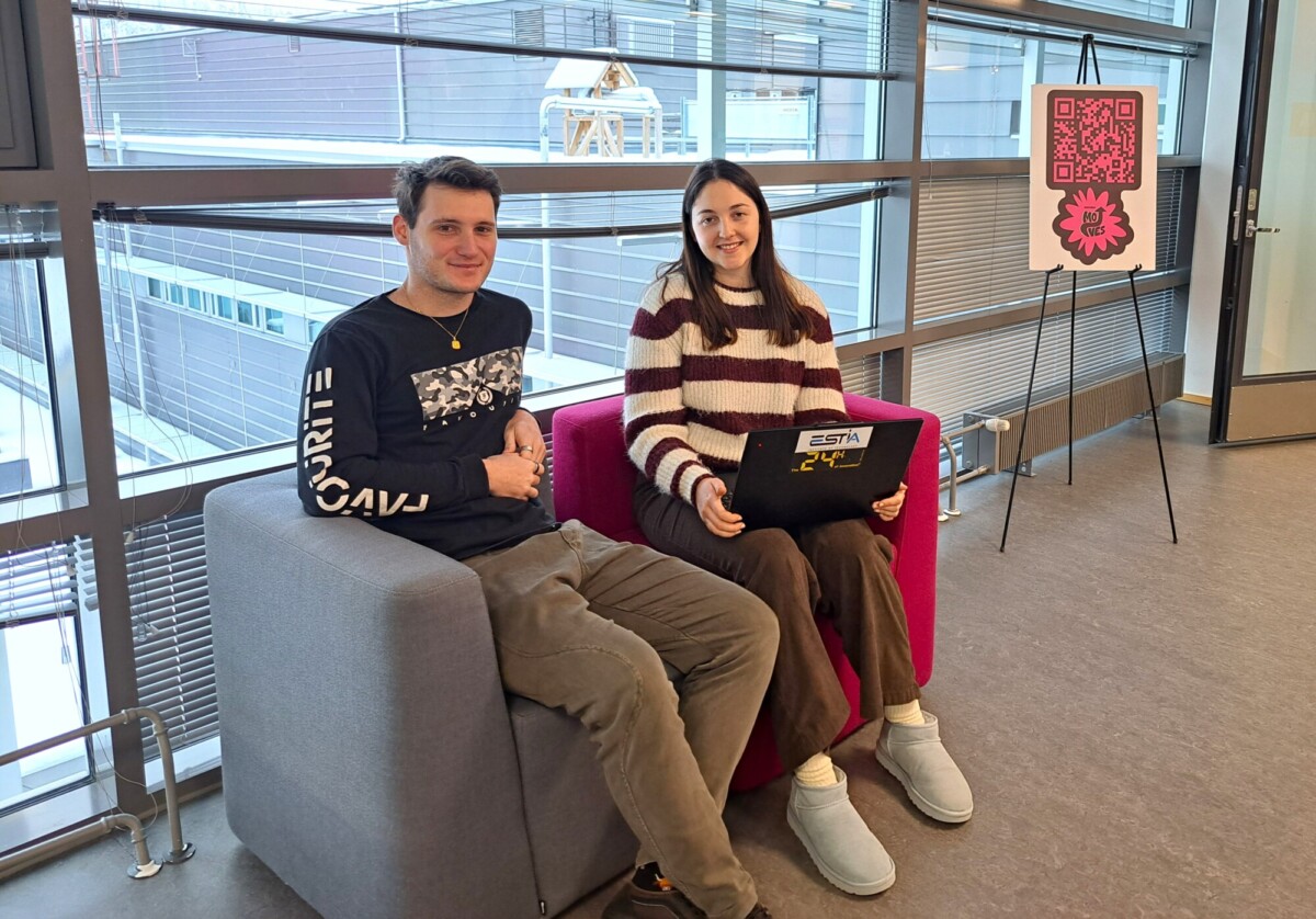 Kaksi ihmistä istuu vierekkäin modernilla nojatuolilla valoisassa huoneessa. Miehellä on musta paita ja harmaat housut. Kannettavaa tietokonetta kädessään pitävällä naisella on raidallinen villapaita ja tossut. Lähellä seisoo kyltti, jossa on vaaleanpunainen QR-koodi.