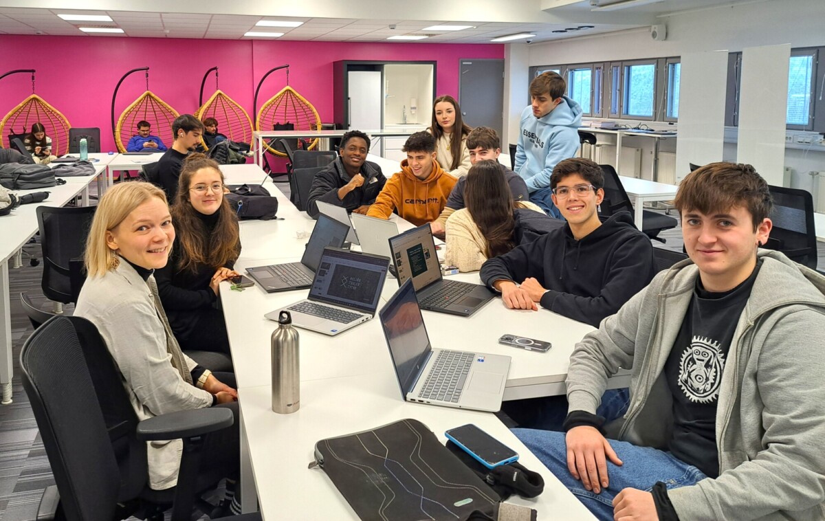 Joukko opiskelijoita istuu valkoisten pöytien ääressä modernissa luokkahuoneessa, jotkut hymyilevät kameralle. Heillä on kannettavat tietokoneet auki, ja taustalla näkyy lisää opiskelijoita vaaleanpunaisten seinien ja riipputuolien lähellä.