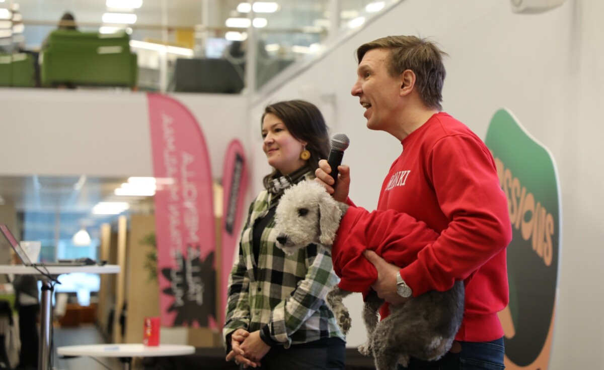 Punapaitainen mies, jolla on koira ja mikrofoni kädessään, puhuu hymyilevän ruudulliseen paitaan pukeutuneen naisen vieressä. He seisovat sisätiloissa kannettavan tietokoneen vieressä, ja taustalla on värikkäitä banderolleja ja toimistokalusteita.