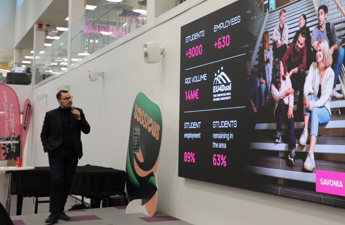 Mustaan pukuun pukeutunut mies puhuu mikrofoniin suuren näytön vieressä, jossa näkyy tilastoja opiskelijoista, työntekijöistä ja projektin volyymista, ja oikealla puolella on otsikko SAVONIA ja ryhmäkuva.