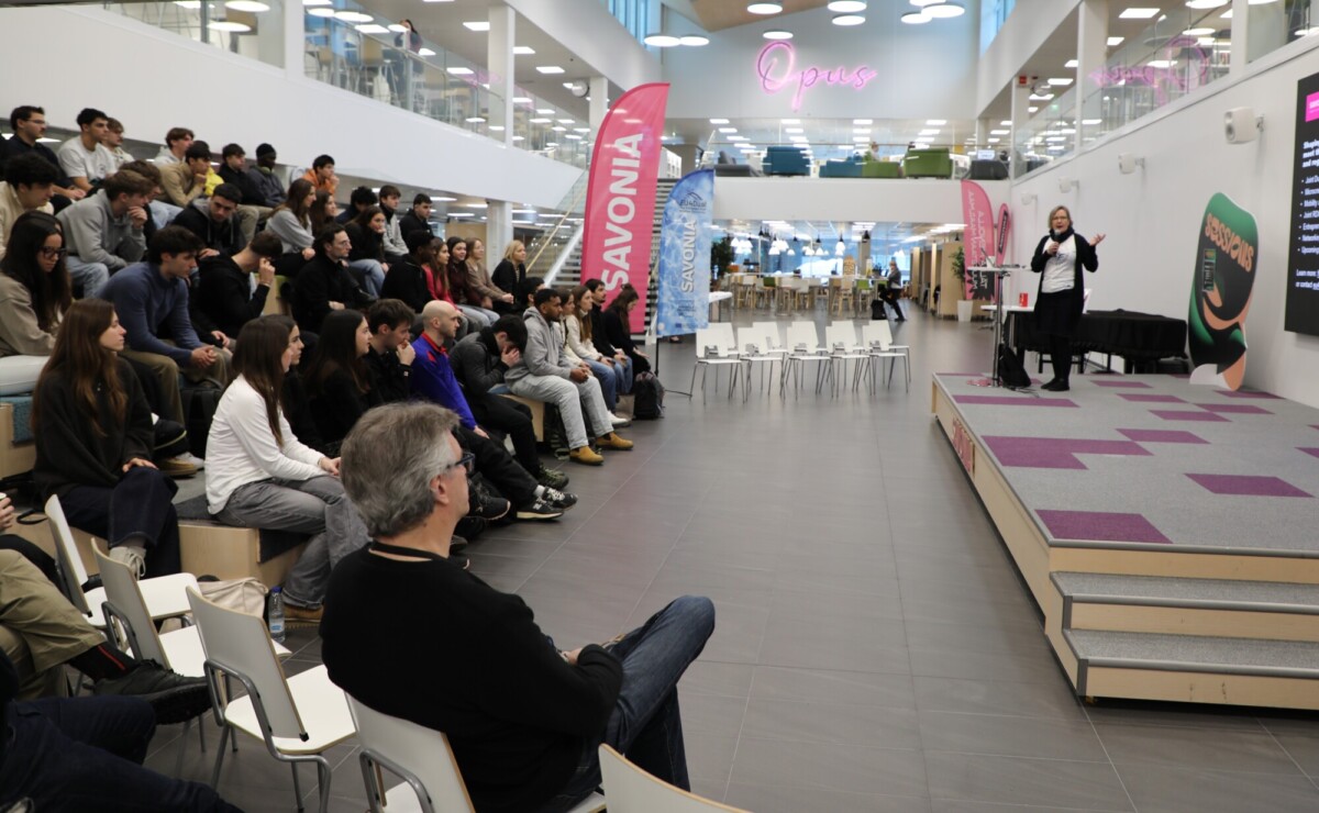 Joukko ihmisiä istuu porrastetuilla penkeillä modernissa, avoimessa tilassa ja kuuntelee tarkkaavaisesti puhujaa, joka esiintyy lavalla vaaleanpunaisten ja sinisten banderollien vieressä. Taustalla näkyy neonvärinen Opus-kyltti.