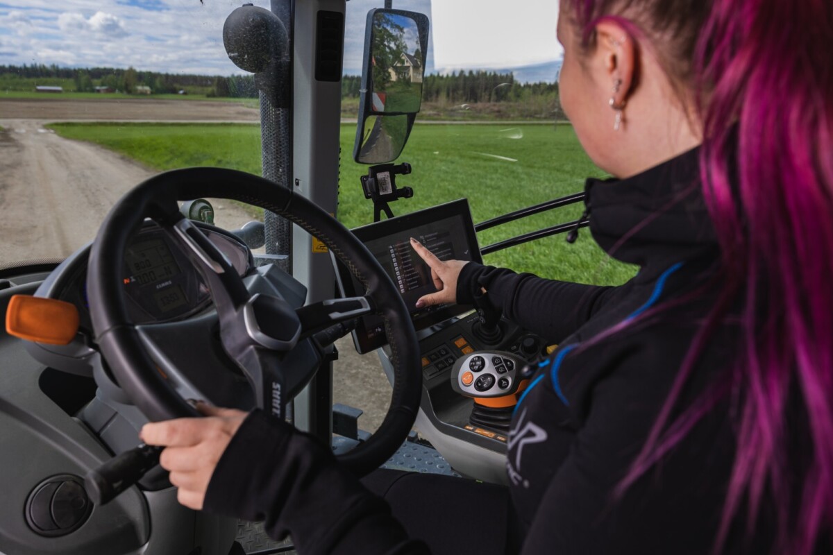Vaaleanpunaiset hiukset omaava henkilö ohjaa nykyaikaista traktoria toisella kädellä ja toisella koskettaa ohjaamossa olevaa digitaalista ohjausnäyttöä, jonka ikkunoista näkyy vihreitä peltoja.