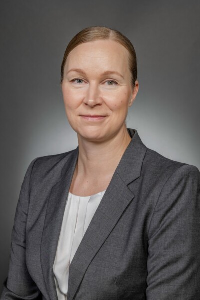 Vaaleaihoinen nainen, jolla on vaaleat, taaksepäin vedetyt vaaleat hiukset ja harmaa bleiseri valkoisen puseron päällä, poseeraa harmaata taustaa vasten ja hymyilee hieman kameralle.