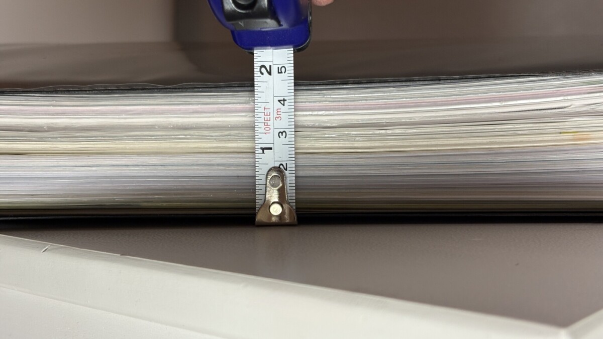 Lähikuva paperipinosta, jota mitataan mittanauhalla, mikä osoittaa, että pino on noin 2 tuumaa paksu. Mittanauha painetaan pystysuoraan arkkia vasten.