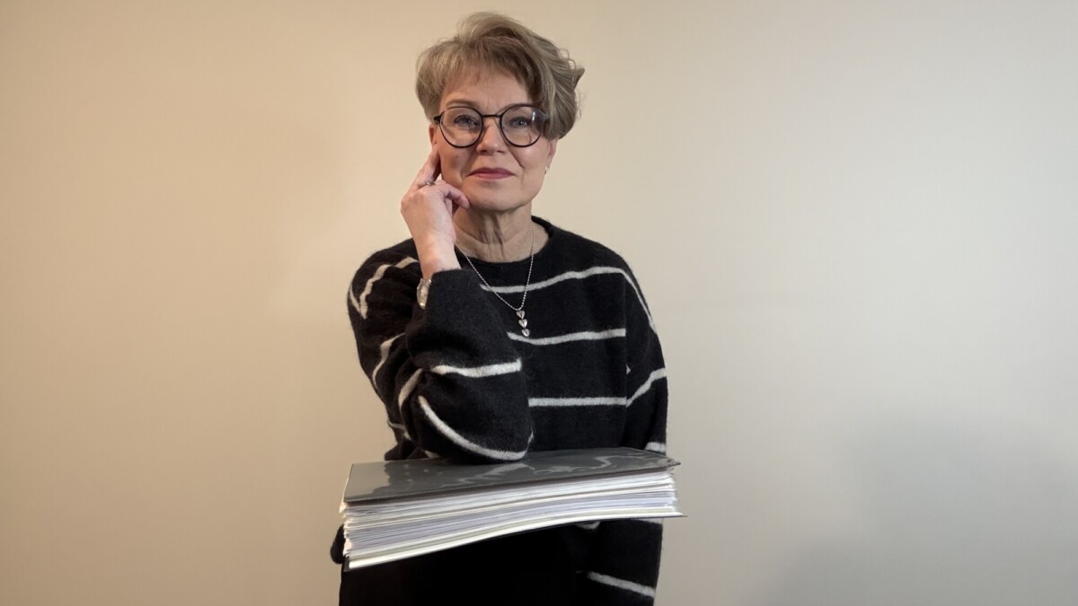 Lyhythiuksinen, silmälasipäinen nainen, jolla on musta, valkoisilla raidoilla varustettu pusero, kädessään pino lehtiä tai papereita ja koskettaa ohimoaan, seisoo vaaleaa taustaa vasten.