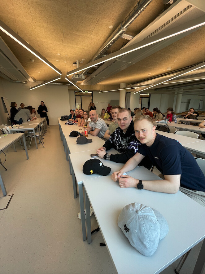Luokkahuone, jossa oppilaat istuvat pöytien ääressä riveissä. Kolme nuorta miestä istuu lähimpänä kameraa ja hymyilee. Pöydillä on baseball-lippalakkeja. Taustalla näkyy lisää oppilaita ja opettaja.