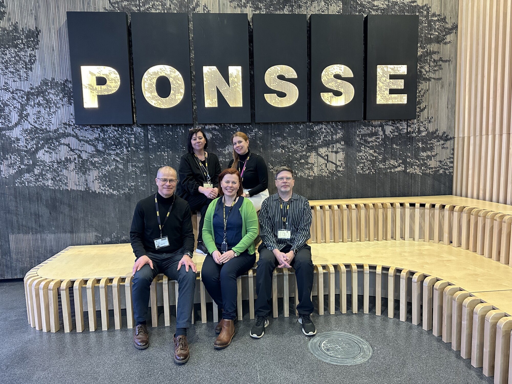 Viisi ihmistä poseeraa ryhmäkuvassa kaarevalla puupenkillä seinän edessä, jossa on suuret valaistut kirjaimet, joissa lukee PONSSE. Taustalla on luonnon inspiroima kuvio.