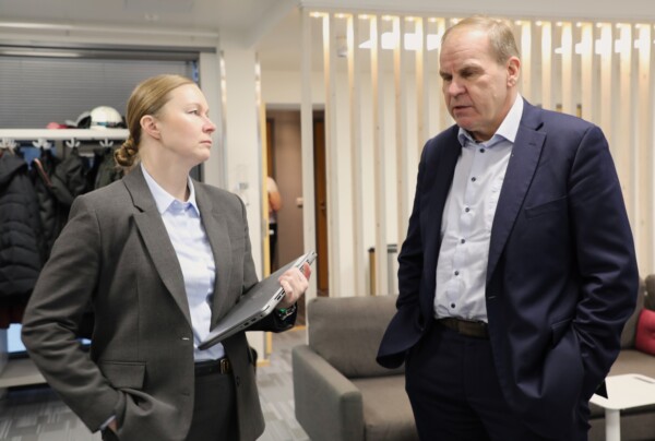 Harmaapukuinen nainen pitää kannettavaa tietokonetta kädessään ja keskustelee tummansiniseen pukuun pukeutuneen miehen kanssa. He seisovat toimistossa, jonka taustalla näkyy sohva ja vaateteline.