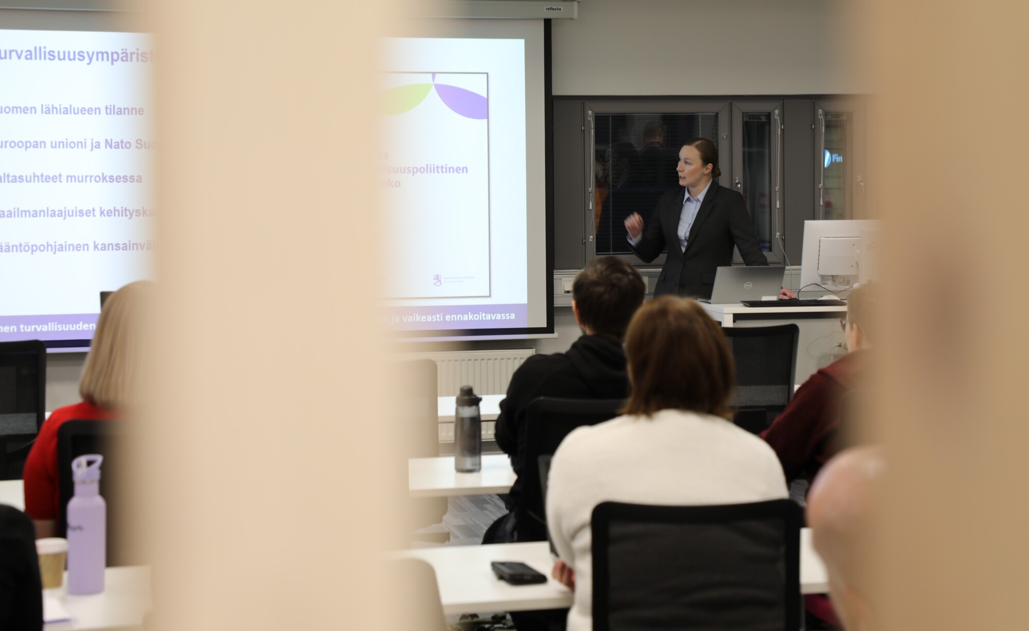 Liikeasuun pukeutunut henkilö seisoo luokkahuoneen edessä ja pitää esitelmää istuville osallistujille. Esitelmöitsijän takana näkyy projisoitu dia, jossa on suomenkielistä tekstiä.