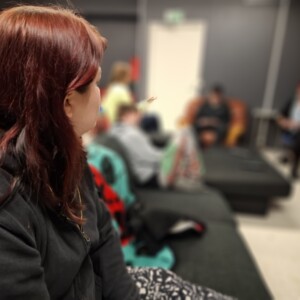 Punatukkainen henkilö istuu etualalla sohvalla ja katsoo kohti ryhmää ihmisiä, jotka istuvat ja keskustelevat epäselvässä taustassa huoneessa, jossa on tummat seinät.