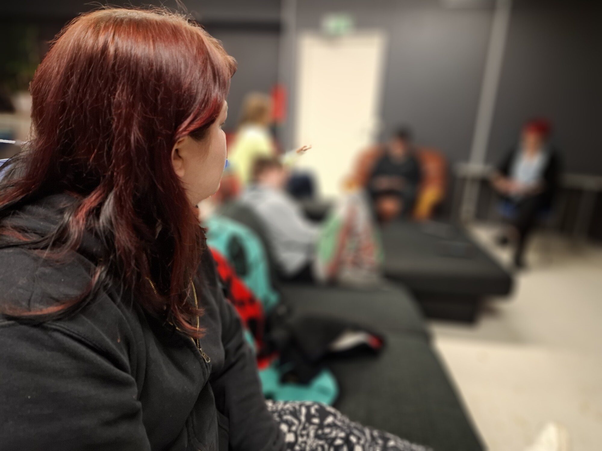 Punatukkainen henkilö istuu etualalla sohvalla ja katsoo kohti ryhmää ihmisiä, jotka istuvat ja keskustelevat epäselvässä taustassa huoneessa, jossa on tummat seinät.