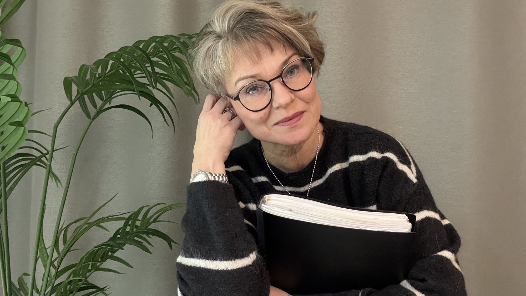 Lyhyet, vaaleat hiukset ja silmälasit omaava nainen hymyilee ja nojaa päätään käteensä. Hänellä on raidallinen pusero ja hänellä on kädessään musta kansio. Taustalla on vihreä kasvi ja harmaa verho.