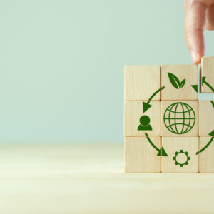 Käsi järjestää puupalikoita, joissa on vihreitä kestävän kehityksen symboleja, kuten maapallo, lehti, henkilö, salama ja vaihde, muodostaen pyöreän kuvion vaalealle puupinnalle.