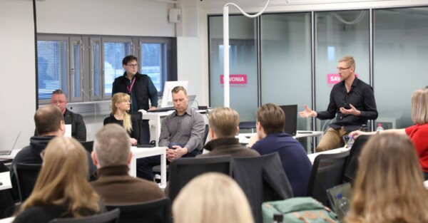 Ryhmä ihmisiä modernissa luokkahuoneessa; yksi mies seisoo ja tekee eleitä, kun muut istuvat ja kuuntelevat, pöydillä on kannettavat tietokoneet ja muistiinpanovälineet ja taustalla suuret ikkunat.