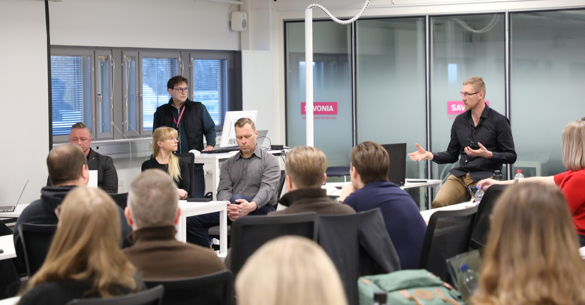 Ryhmä ihmisiä modernissa luokkahuoneessa; yksi mies seisoo ja tekee eleitä, kun muut istuvat ja kuuntelevat, pöydillä on kannettavat tietokoneet ja muistiinpanovälineet ja taustalla suuret ikkunat.