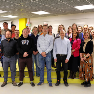 Noin kahdenkymmenenviiden aikuisen ryhmä, sekä miehiä että naisia, seisoo yhdessä kirkkaasti valaistussa sisätilassa, poseeraa ja hymyilee ryhmäkuvaa varten valkoisen seinän ja projektorinäytön edessä.