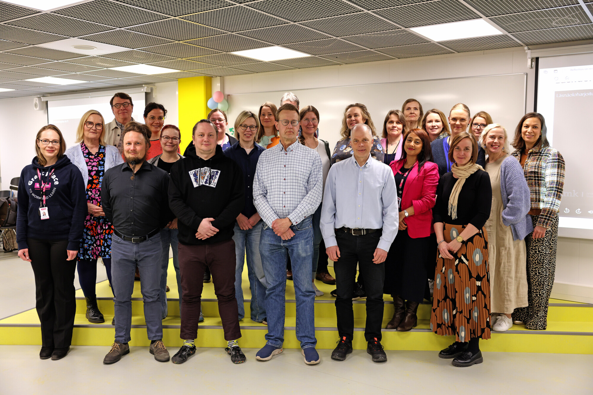 Noin kahdenkymmenenviiden aikuisen ryhmä, sekä miehiä että naisia, seisoo yhdessä kirkkaasti valaistussa sisätilassa, poseeraa ja hymyilee ryhmäkuvaa varten valkoisen seinän ja projektorinäytön edessä.