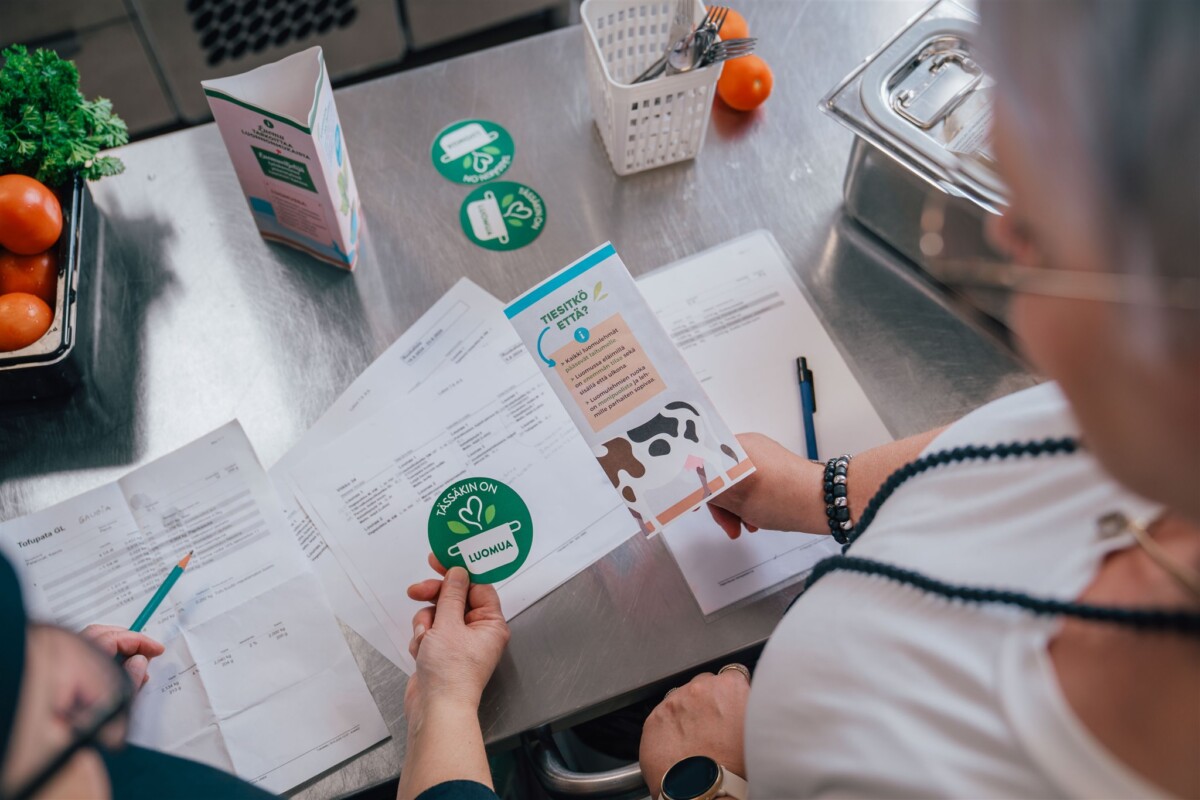 Kaksi ihmistä tarkastelee tulostettuja asiakirjoja ja vihreitä elintarvikesertifiointitarroja ruostumattomasta teräksestä valmistetulla pöydällä, jota ympäröivät tuoreet tomaatit, lehtivihannekset, kynä ja muovisäiliö.