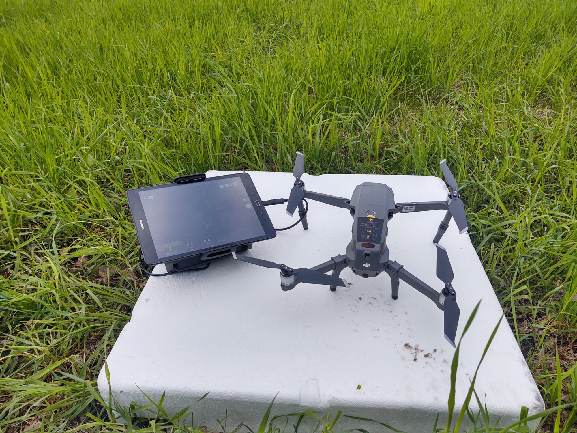 Neljällä potkurilla varustettu lennokki, johon on asennettu kamera, istuu valkoisella alustalla nurmikentällä ohjaimeen kiinnitetyn tabletin vieressä.