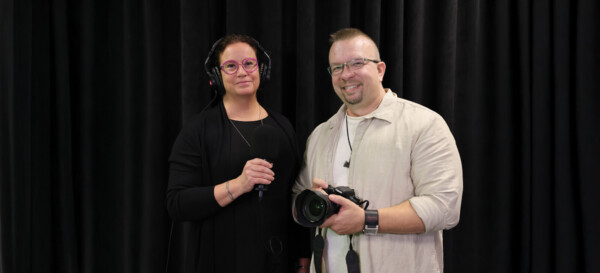 Kaksi ihmistä seisoo mustan verhon edessä; vasemmalla olevalla naisella on kuulokkeet ja hänellä on mikrofoni, kun taas oikealla oleva mies hymyilee ja pitää kameraa.