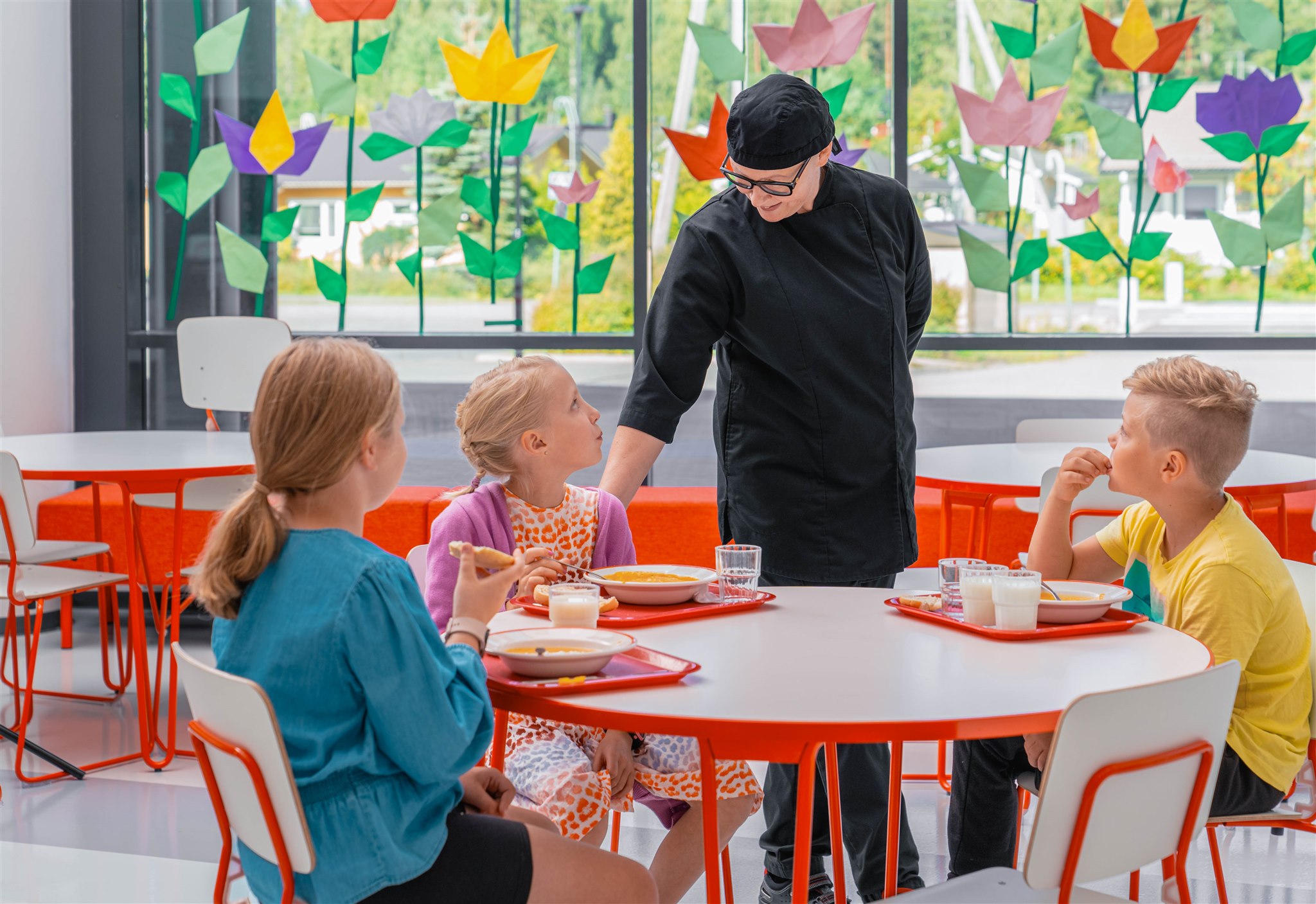 Mustaan univormuun pukeutunut kahvilatyöntekijä puhuu kolmen lapsen kanssa, jotka istuvat pyöreän pöydän ääressä syömässä lounasta. Värikkäät paperikukat koristavat valoisan, modernin ruokailutilan suuria ikkunoita.