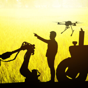 Siluetti henkilöstä, joka osoittaa robottikättä, yläpuolella lentää lennokki ja vieressä on traktori kirkkaalla pellolla, joka symboloi nykyaikaista teknologiaa maataloudessa.