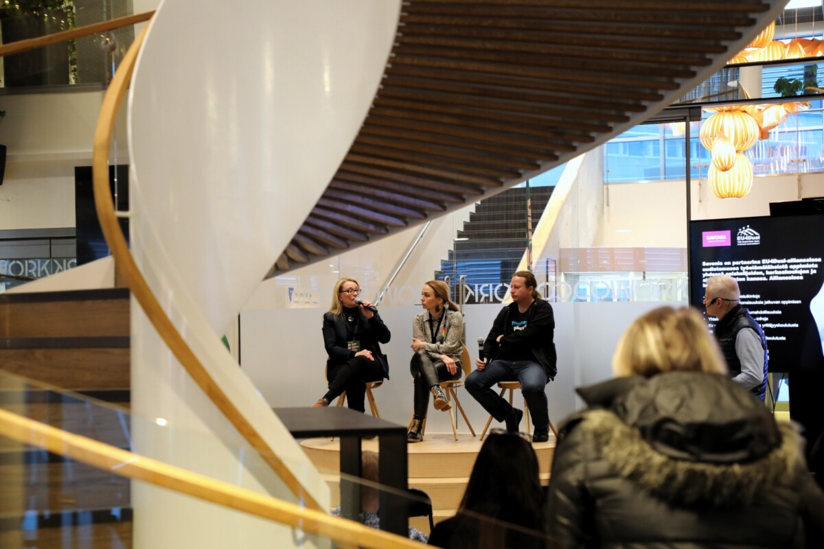 Neljä ihmistä istuu pienellä näyttämöllä paneelikeskustelussa. Kohtausta kehystää etualalla oleva kaareva portaikko, jota yleisö seuraa. Taustalla näkyy moderni sisustus ja suuret ikkunat.