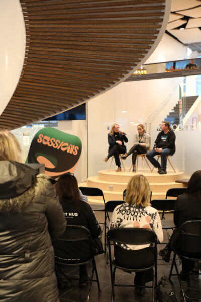 Kolme ihmistä istuu pienellä näyttämöllä modernissa sisätilassa ja puhuu mustilla tuoleilla istuvalle yleisölle. Vasemmalla on kyltti, jossa lukee Sessions. Taustalla näkyy kierreportaat ja lasiseinät.