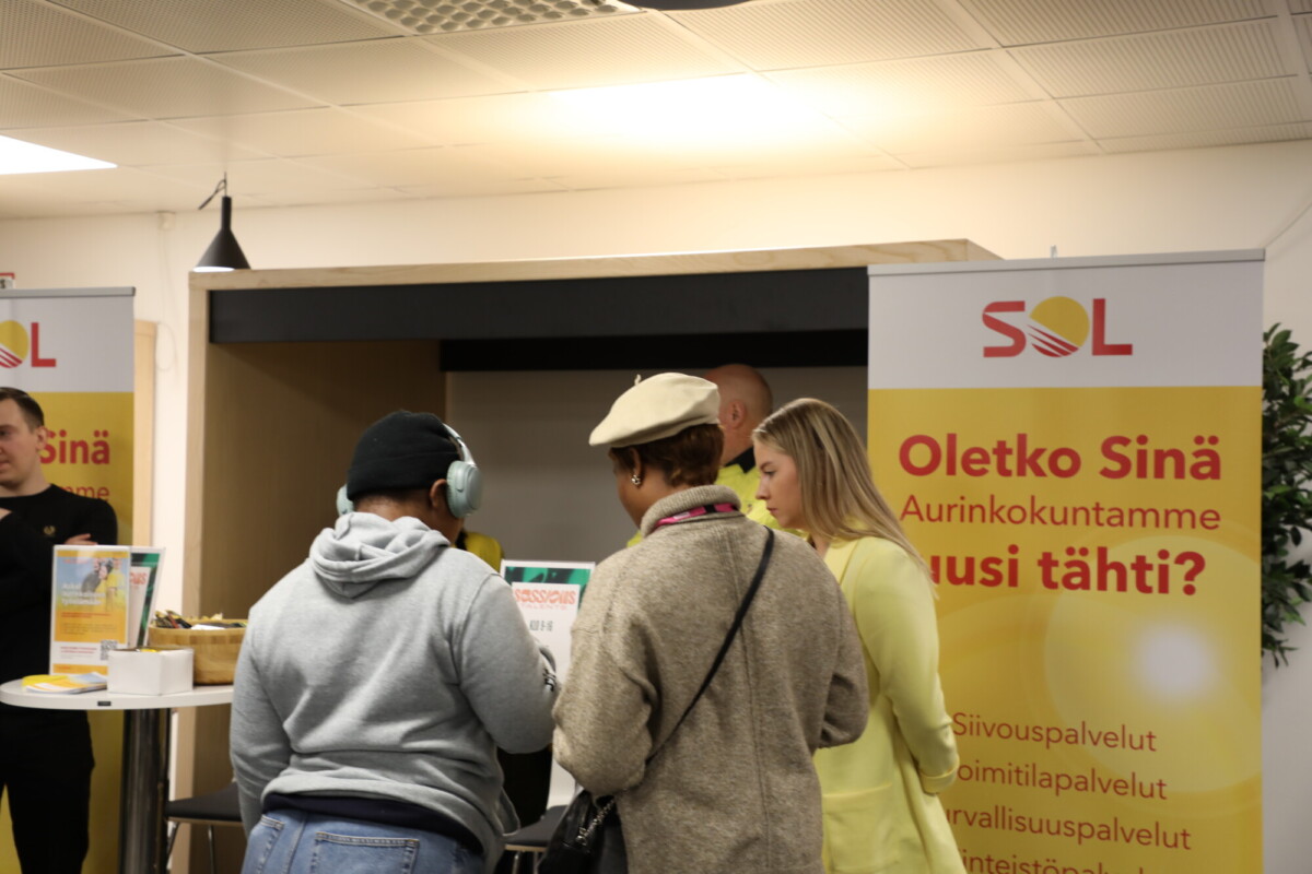 Kolme ihmistä seisoo kojulla, jossa on SOLin suomenkieliset banderollit. Yhdellä henkilöllä on kuulokkeet, toisella beige baretti ja kolmannella keltainen takki. Ympäristö näyttää olevan sisätiloissa järjestettävä tapahtuma tai työpaikkamessut.