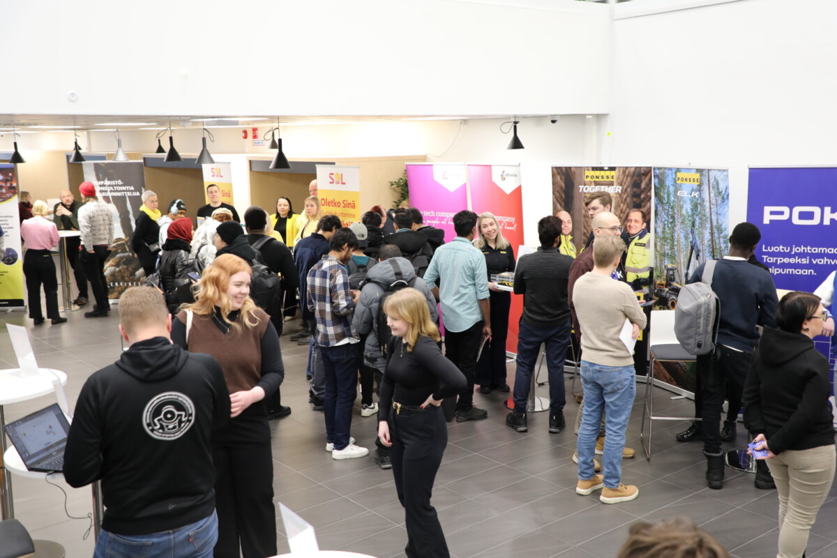 Suuri joukko ihmisiä on vuorovaikutuksessa sisätiloissa järjestettävillä työnhakumessuilla tai näyttelyssä, vierailee eri kojuilla, joissa on banderolleja, ja keskustelee edustajien kanssa. Ympäristö on valoisa, moderni ja kiireinen.