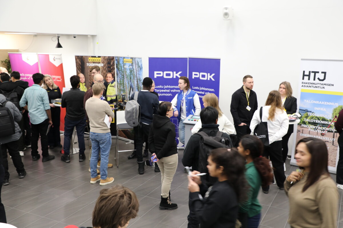 Ihmiset ovat vuorovaikutuksessa uramessuilla tai verkostoitumistapahtumissa tiedotuskojujen ja banderollien kanssa. Useat ryhmät keskustelevat, kun taas toiset kävelevät ohi tai katselevat näyttöjä valoisassa sisätilassa.