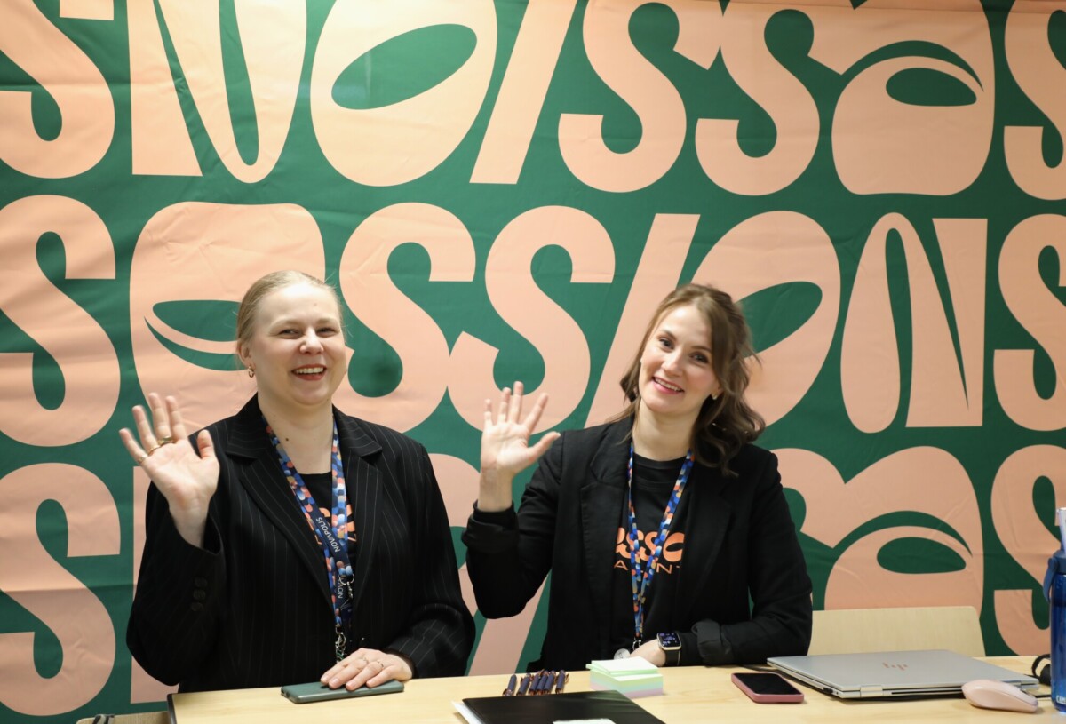 Kaksi naista istuu pöydän ääressä, hymyilee ja vilkuttaa kameralle. Heillä on yllään mustat bleiserit ja kaulanauhat, ja taustalla on vihreä ja persikkainen SESSIONS-banneri.