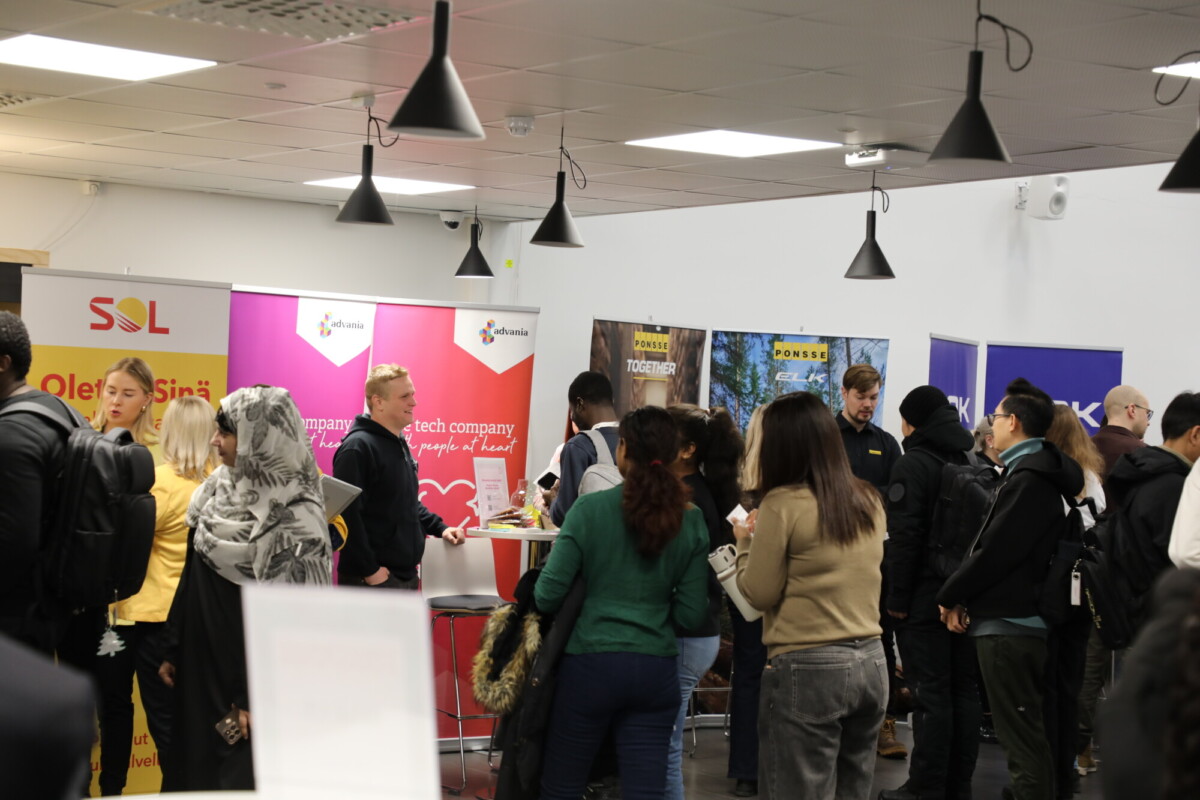 Joukko ihmisiä on vuorovaikutuksessa eri yritysten osastoilla sisätiloissa järjestettävien uramessujen tai verkostoitumistapahtuman aikana. Bannerit ja tiedotusmateriaalit ovat näkyvillä, ja osallistujat keskustelevat.